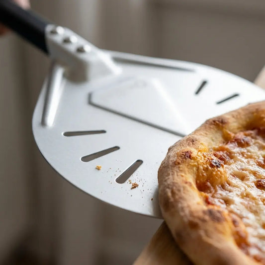 Gros plan de la petite tête ronde perforée glissant sous le bord d’une pizza pour la faire pivoter sur la pierre de cuisson.