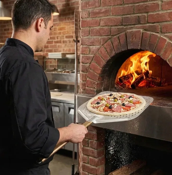 Un homme en veste noire tenant une pelle perforée avec une pizza Margherita prête à l'enfournement devant un four à bois en brique"