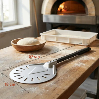 Petite pelle de rotation perforée posée sur une table en bois devant un four à pizza allumé en arrière‑plan, suggérant un usage régulier. Taille 57 cm sur 18 cm.