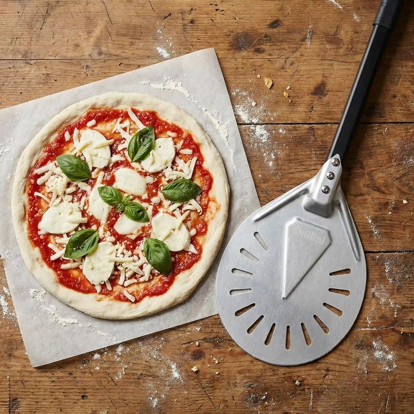 Pelle de rotation perforée posée sur un plan de travail en bois à côté d’une pizza margherita, dans un environnement de préparation de pizzas.