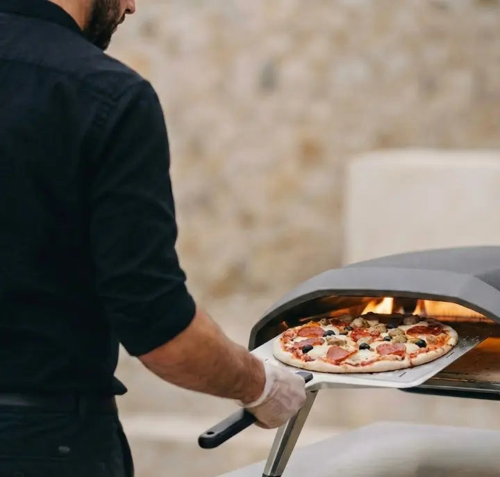 Pizzaiolo enfournant une pizza dans un mini four ooni, avec sa pelle à pizza perforée