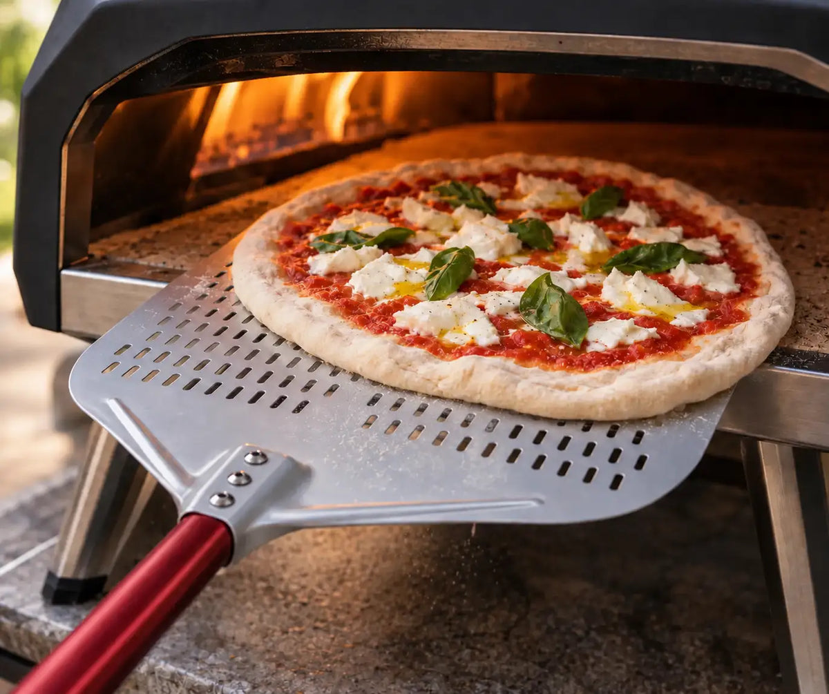 Pelle à pizza perforée en aluminium avec manche rouge, utilisée pour enfourner une pizza dans un four à pizza