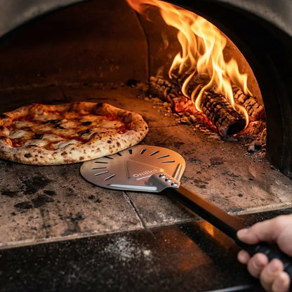 Main d’un pizzaiolo utilisant une pelle de rotation perforée à petite tête pour tourner une pizza dans un four à pizza à flamme vive.
